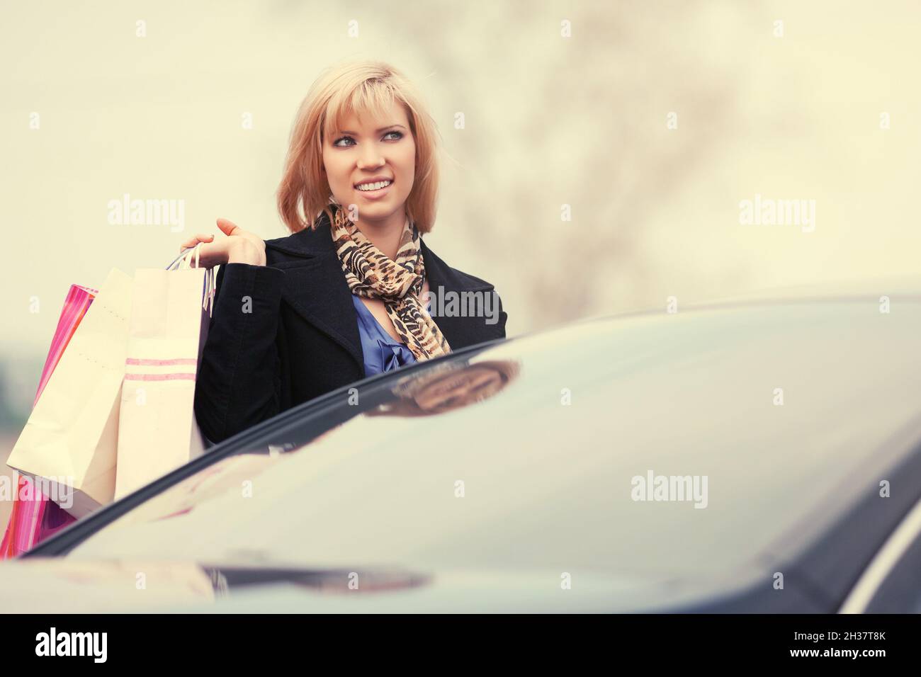Felice giovane donna di moda con borse per la spesa accanto a lei Auto elegante modello femminile in classico camice nero e seta sciarpa Foto Stock