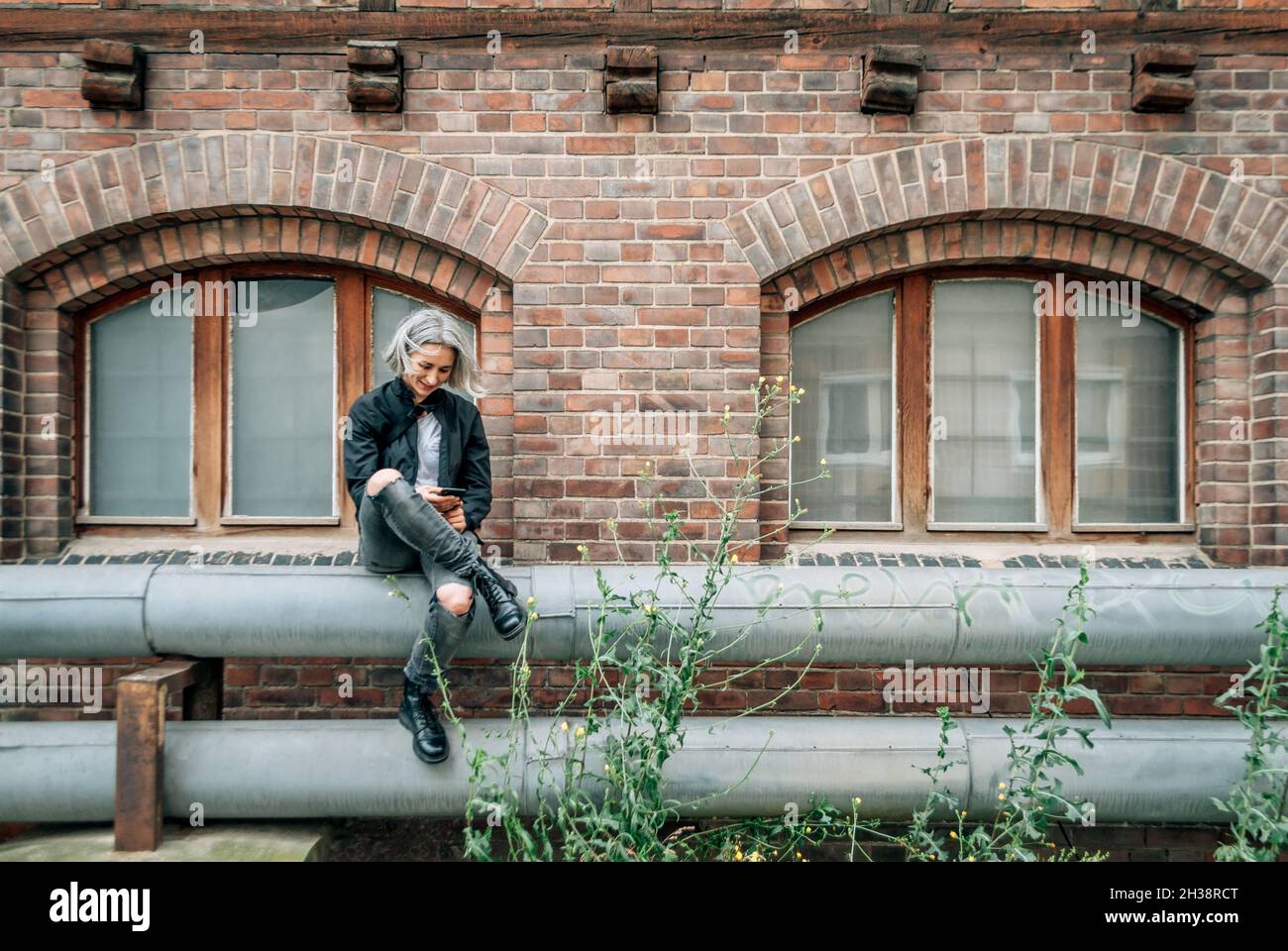 La donna si siede sul tubo vicino a un edificio abbandonato, guarda lo smartphone Foto Stock
