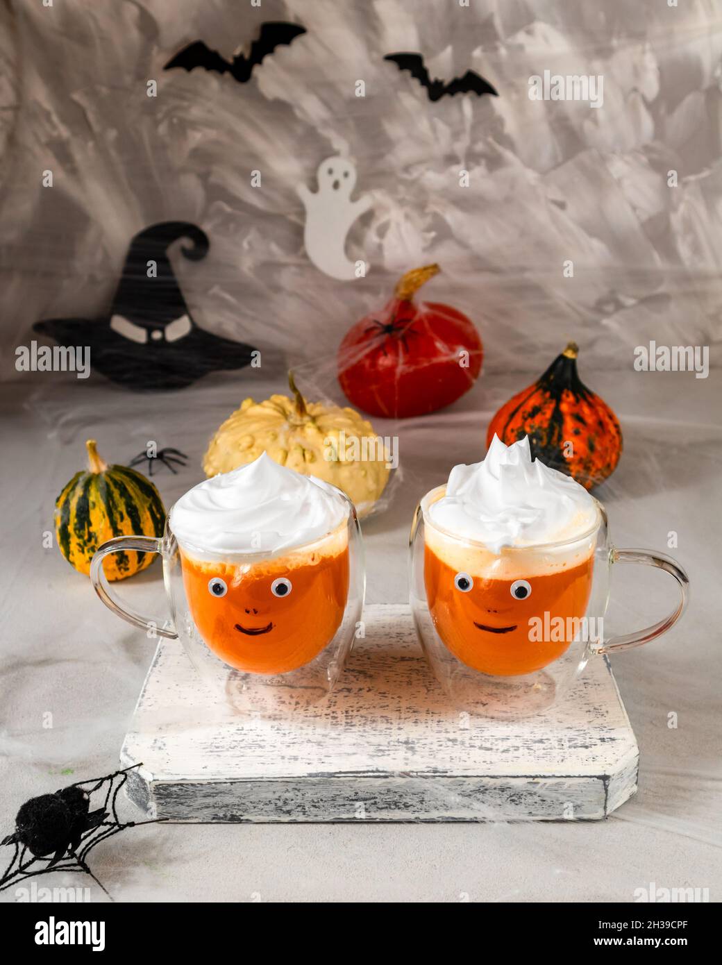 Due tazze di vetro di succo di zucca con facce dipinte sullo sfondo delle zucche di Halloween su una parete grigia con fantasmi, pipistrelli, ragni, cappelli di strega Foto Stock
