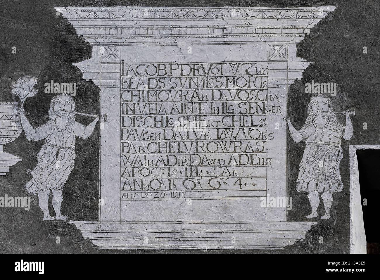 I sostenitori femminili soffiano corna e tengono fiori in alto come fiancheggiano un'iscrizione in lingua romanica datata 1664, in affascinante arte sgraffito rustico sulla facciata di una casa tradizionale Engadiner in Ardez, Graubünden o Grigioni cantone, Svizzera orientale. I residenti di Ardez sono tra circa 40,000 cittadini svizzeri la cui prima lingua è il Romansh, una lingua legacy dell'antico impero romano. Foto Stock