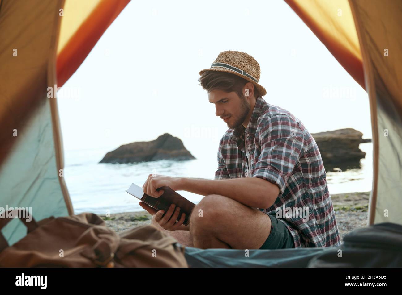 Un ragazzo europeo ha letto un libro sulla spiaggia di sabbia. Vista dalla tenda. Giovane uomo concentrato. Concetto di campeggio vacanza sulla natura. Turismo estivo e traversata Foto Stock