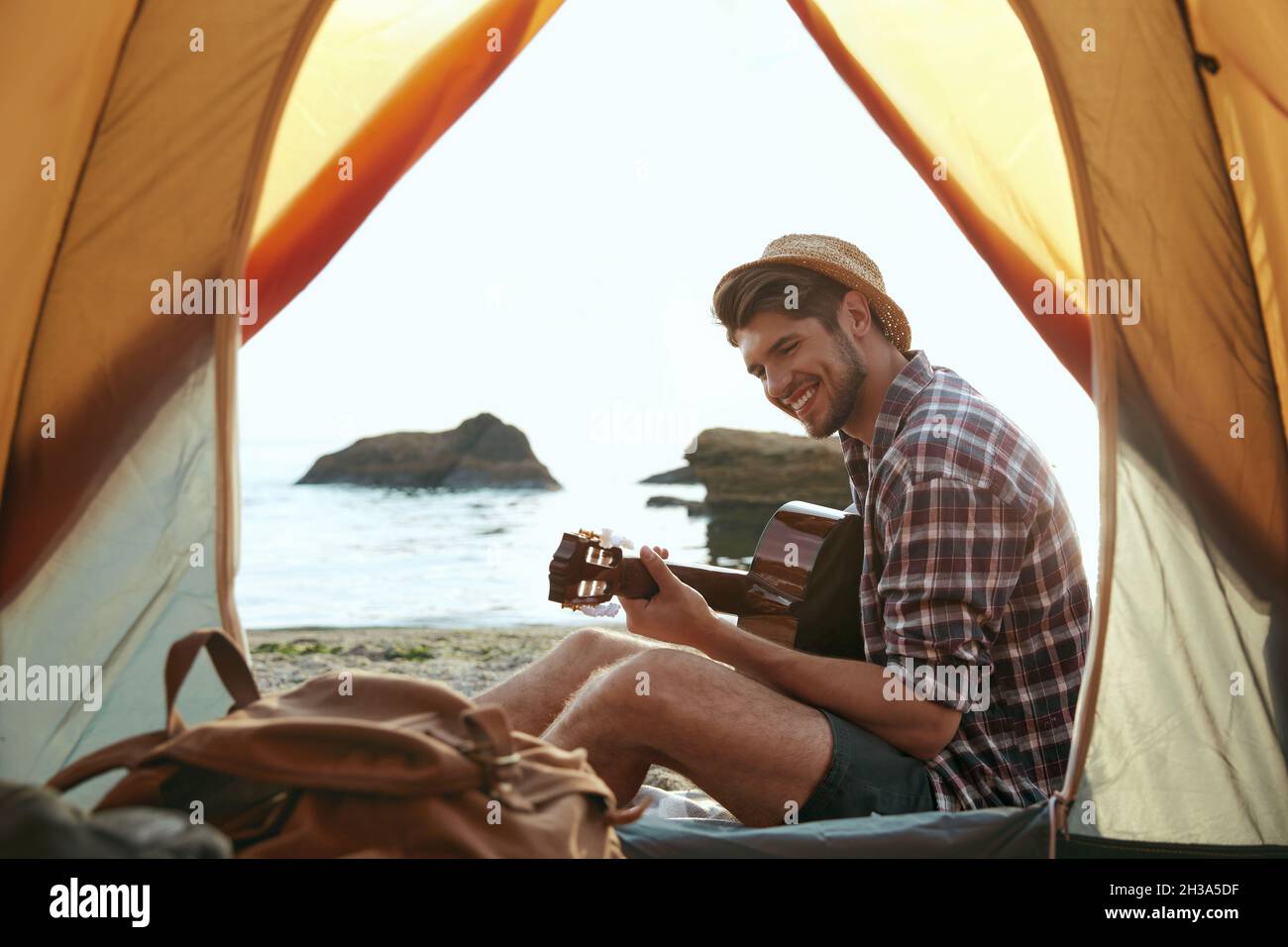 Il ragazzo europeo suona la chitarra sulla spiaggia sabbiosa del mare. Vista dalla tenda. Giovane uomo sorridente. Concetto di campeggio vacanza sulla natura. Turismo estivo e travelli Foto Stock