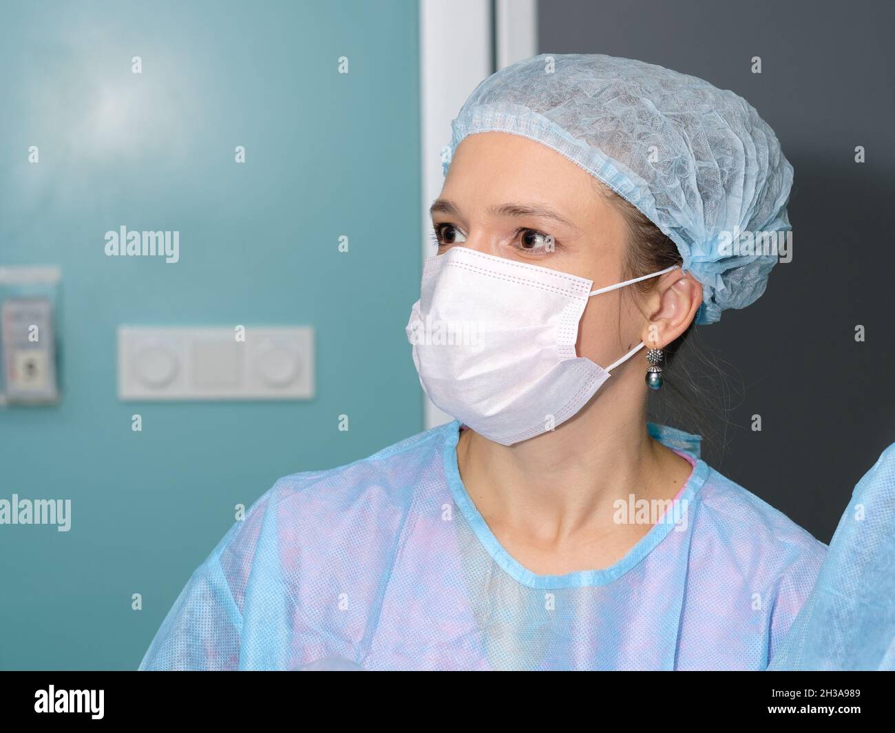 Giovane medico femminile in uniforme blu con cappuccio chirurgico e maschera di protezione, ritratto da vicino. Prevenzione di Covid-19. Sguardo vicino di occhi marroni di medico femminile in uniforme medica. Foto Stock