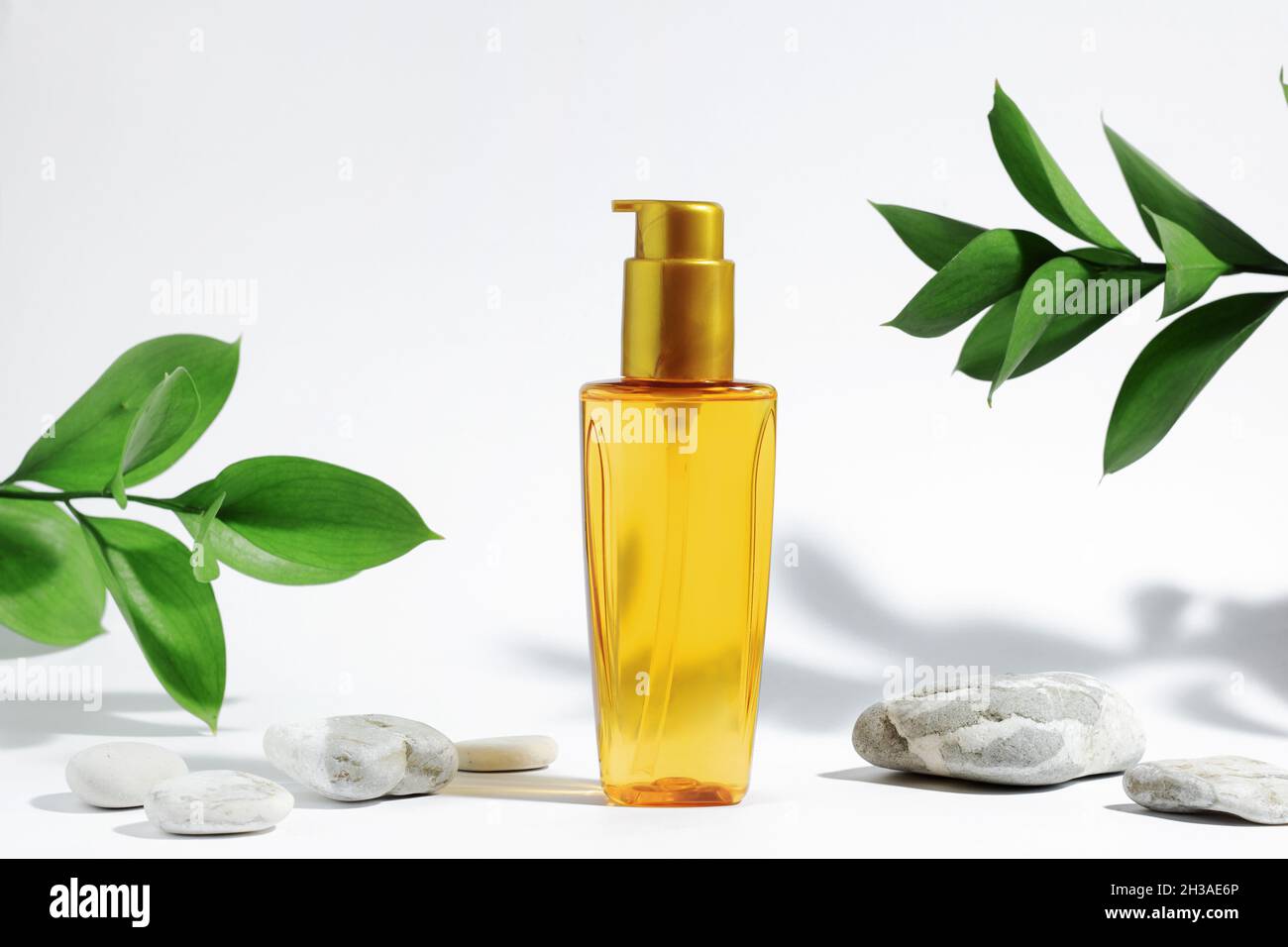 Bottiglia trasparente di olio arancione giallo per proteggere il sole, abbronzarsi o i capelli. Sfondo bianco con ombre, piante e pietre dure. Cosmetici per la cura estiva. Foto Stock