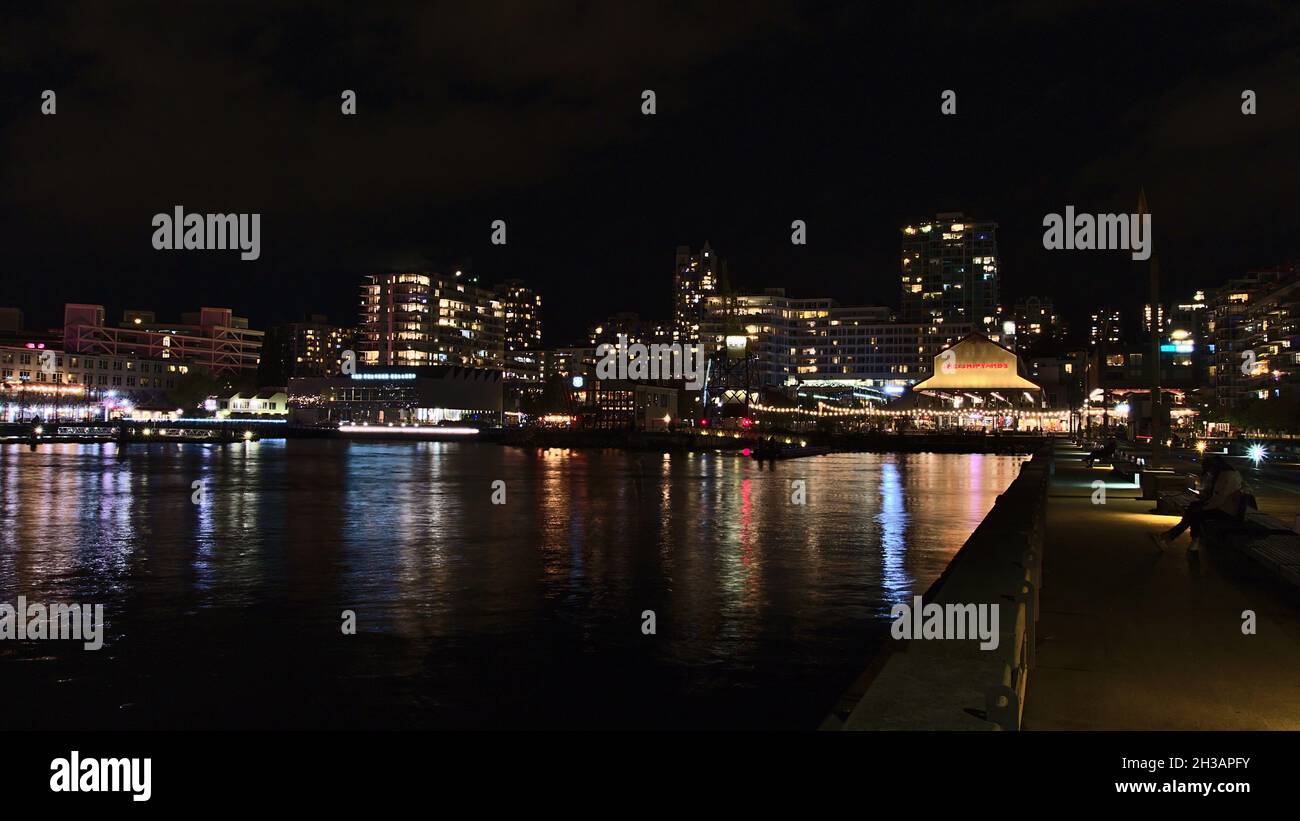 Splendida vista notturna dei cantieri navali, un quartiere a North Vancouver, con persone che si rilassano sul molo e gli edifici illuminati. Foto Stock