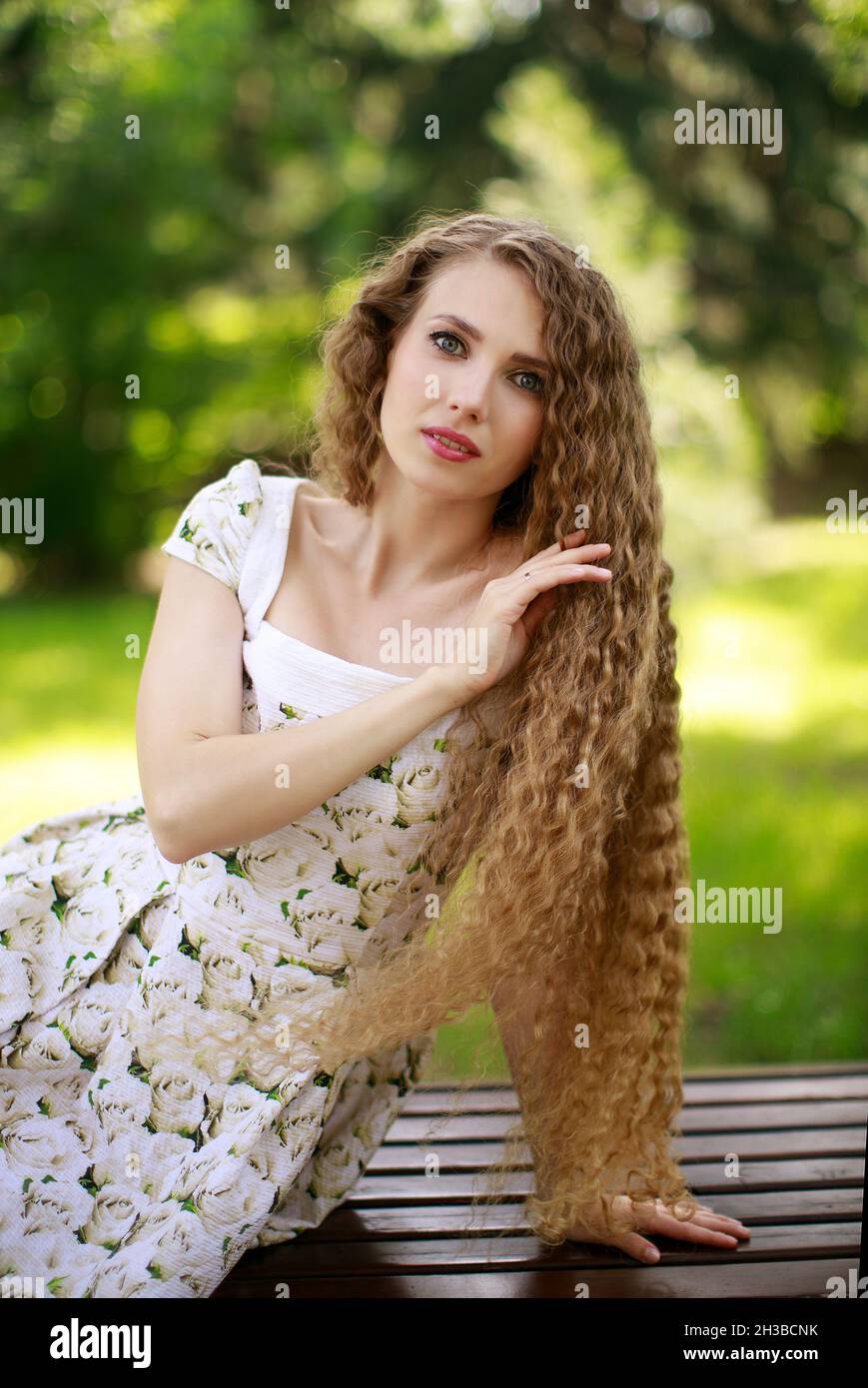 Giovane bella ragazza bionda con capelli lunghi in un abito bianco estivo in posa mentre si siede su una panca parco Foto Stock