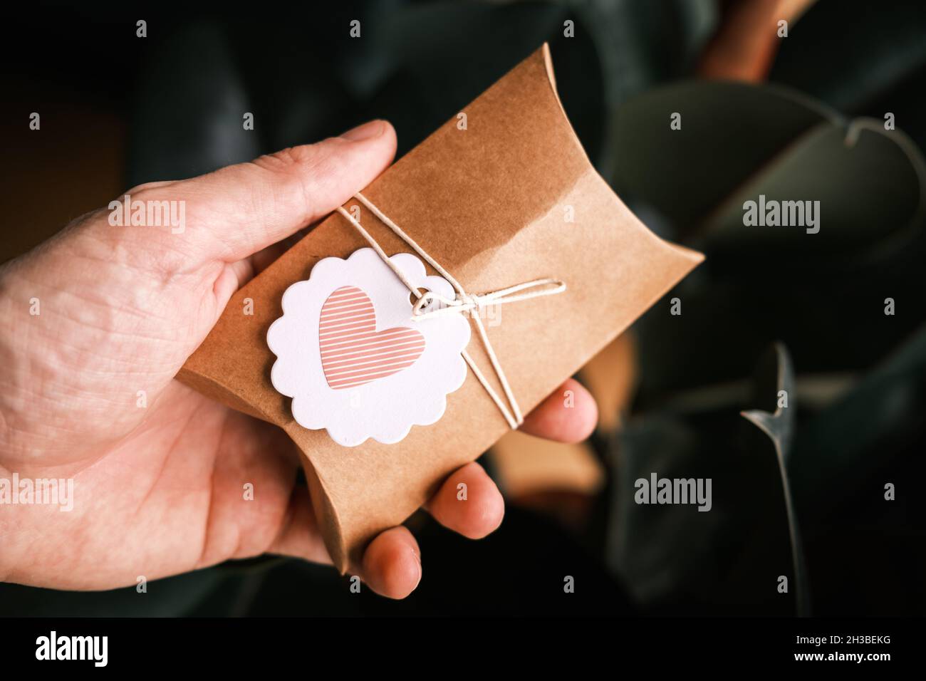 Maschio mano che tiene piccolo regalo avvolto in carta artigianale su sfondo verde scuro. Regalo fatto in casa, forma del cuore sull'etichetta del regalo. Confezione regalo sostenibile. Foto Stock