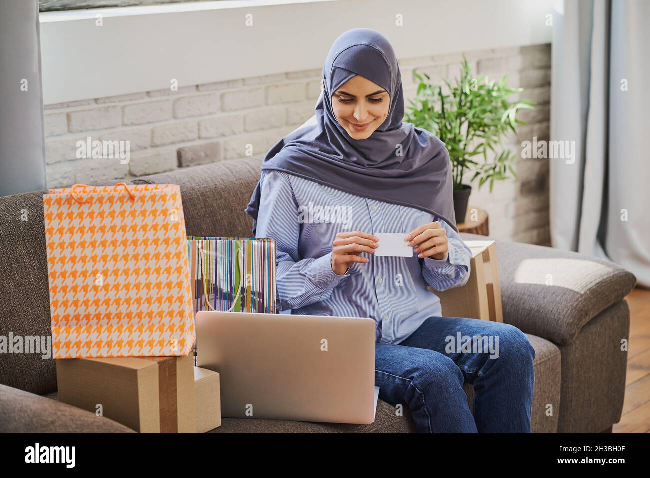 Donna musulmana graziosa seduta su un divano e comprare qualcosa in linea Foto Stock