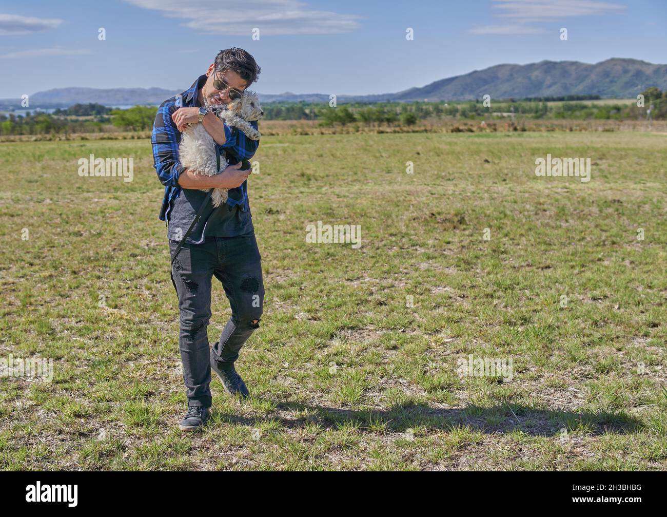 Latino brunette ragazzo in stile urbano abbracciando il suo puppy giocattolo barbolo in campo. Amicizia. Orizzontale. Montagne sfocate sullo sfondo. Vacanze ad Argenti Foto Stock