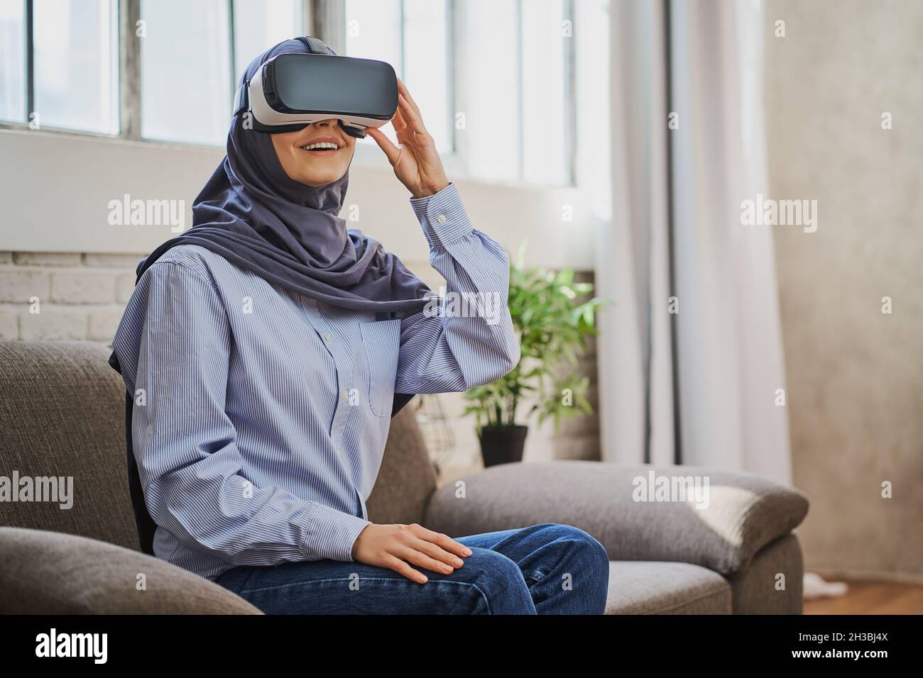Gioiosa donna araba seduta su un divano, con un casco VR e sorridente Foto Stock
