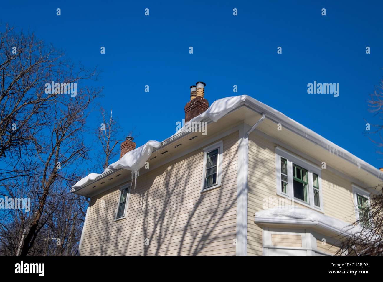 L'angolo esterno di una casa di legno giallo con finestre, un camino, e finiture bianche. C'è neve sul tetto dell'edificio sospeso sopra la vigilia Foto Stock