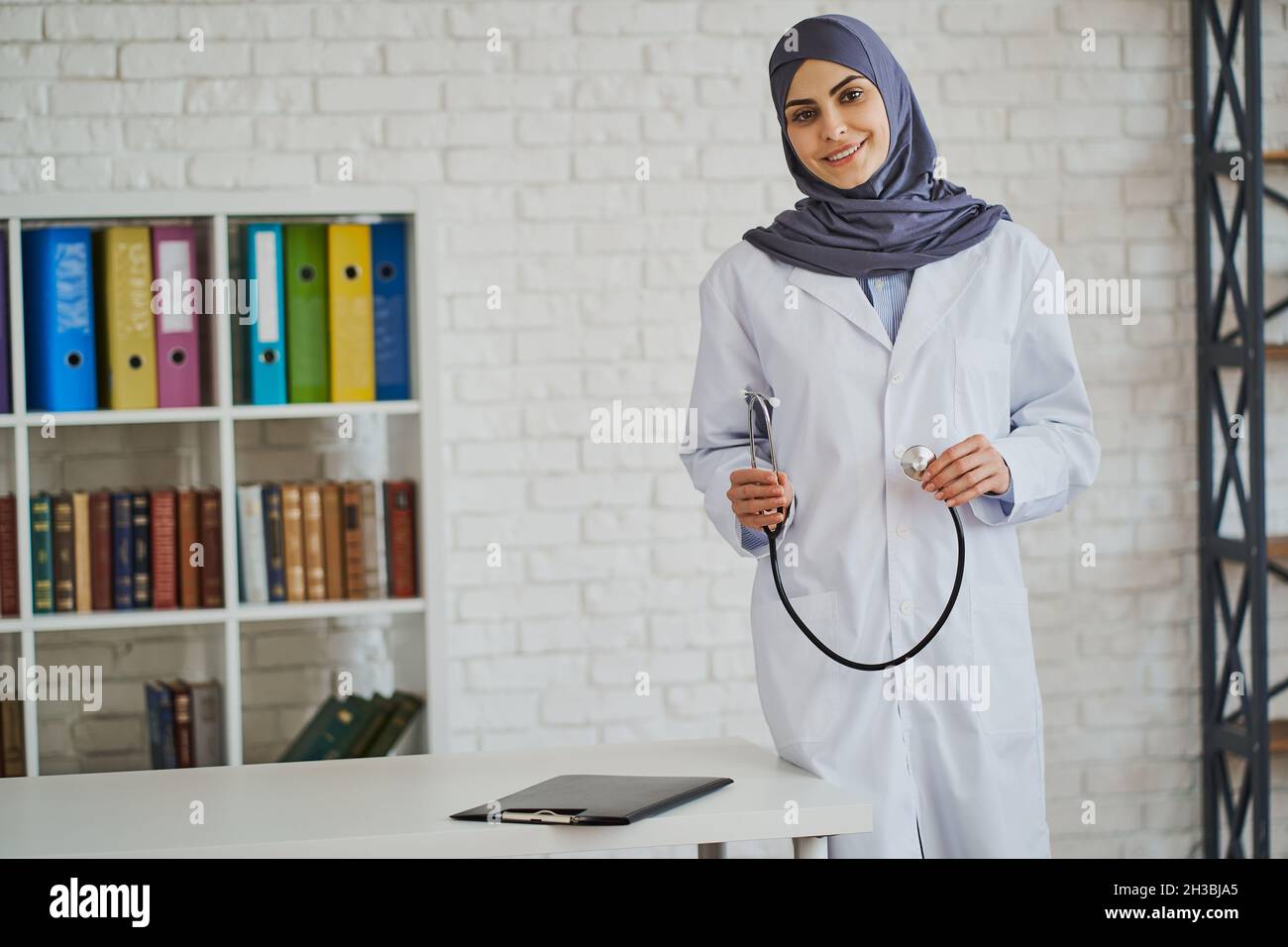 Medico musulmano amichevole che posa con uno stetoscopio vicino al suo posto di lavoro Foto Stock