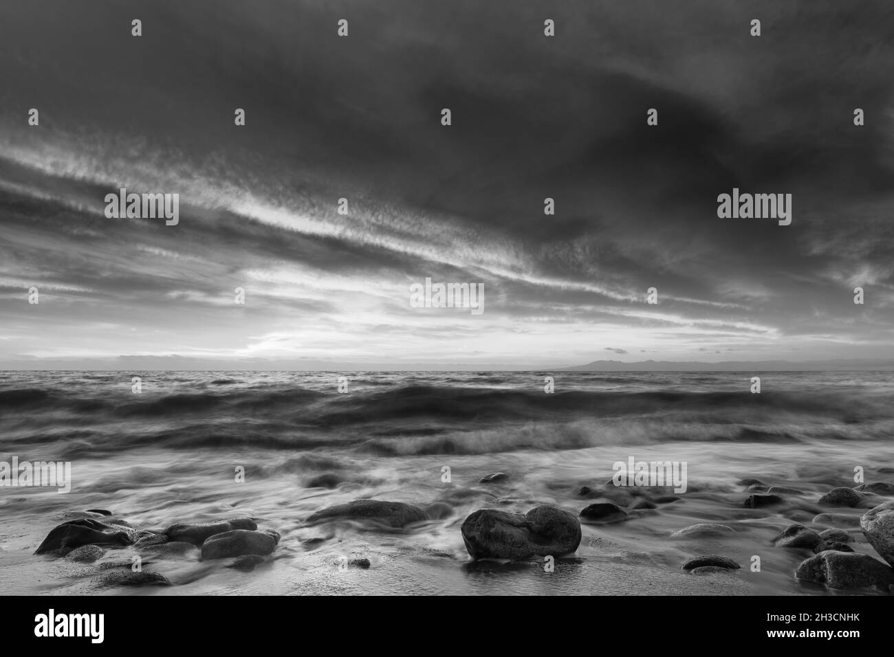 Un Ocean Seascape Sunset Sky con un dettagliato Moody Cloudscape come una dolce onda rotola a Shore in bianco e nero immagine formato Foto Stock