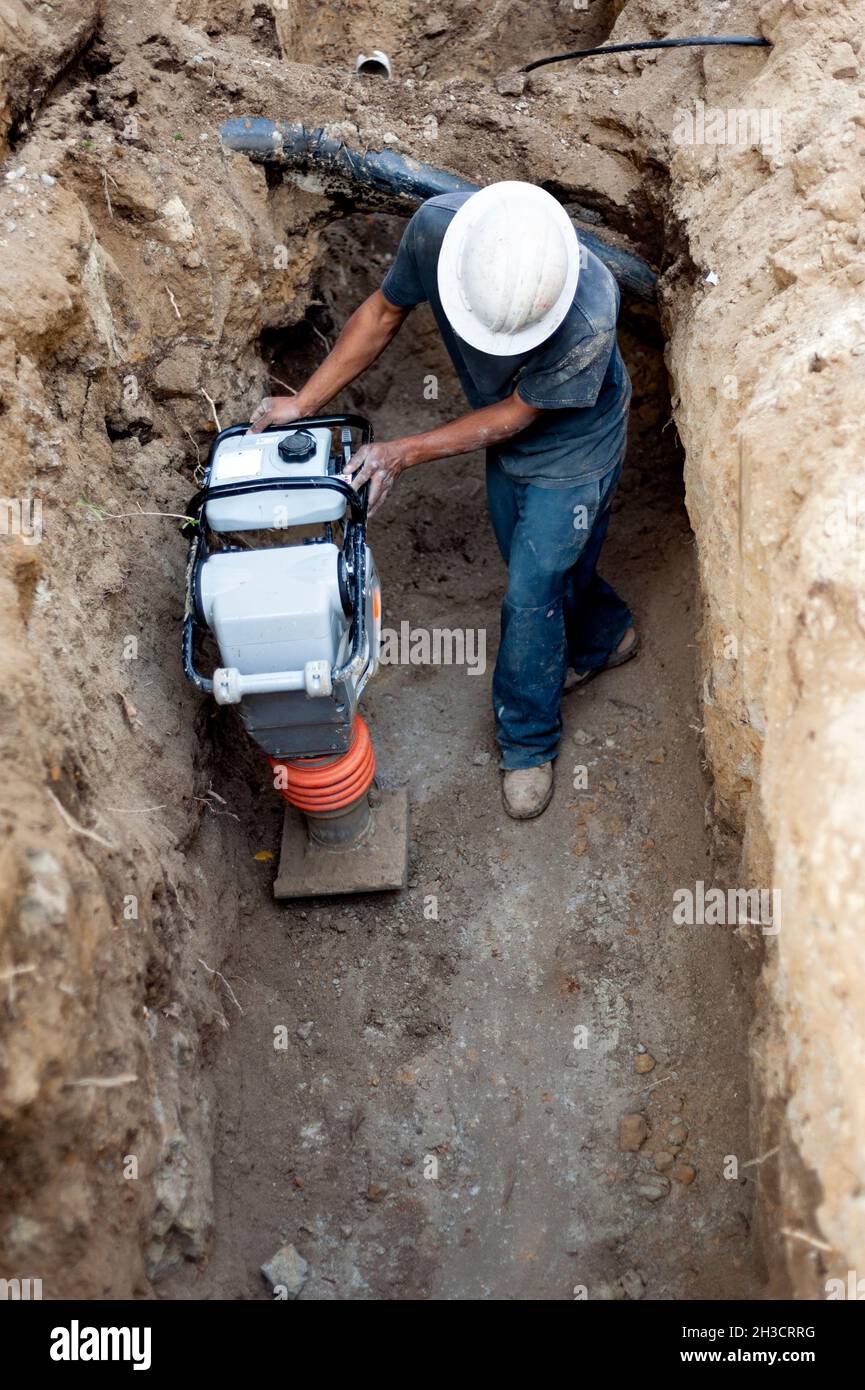 Operaio di costruzione che opera un compattatore di suolo in una trincea profonda Foto Stock