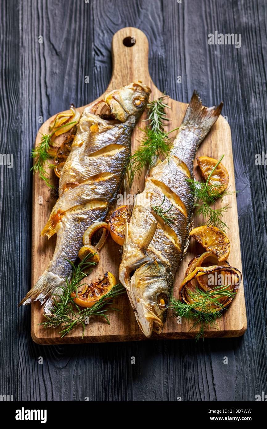 Pesce di branzino arrosto intero servito su un asse di legno con sale marino, aneto, limone su un tavolo di legno nero, vista dall'alto, primo piano Foto Stock