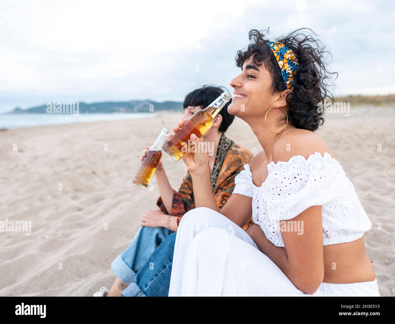 Positivo multietnico femmina migliori amici bere birra fredda mentre si siede sulla spiaggia sabbiosa e ridendo in estate sera Foto Stock