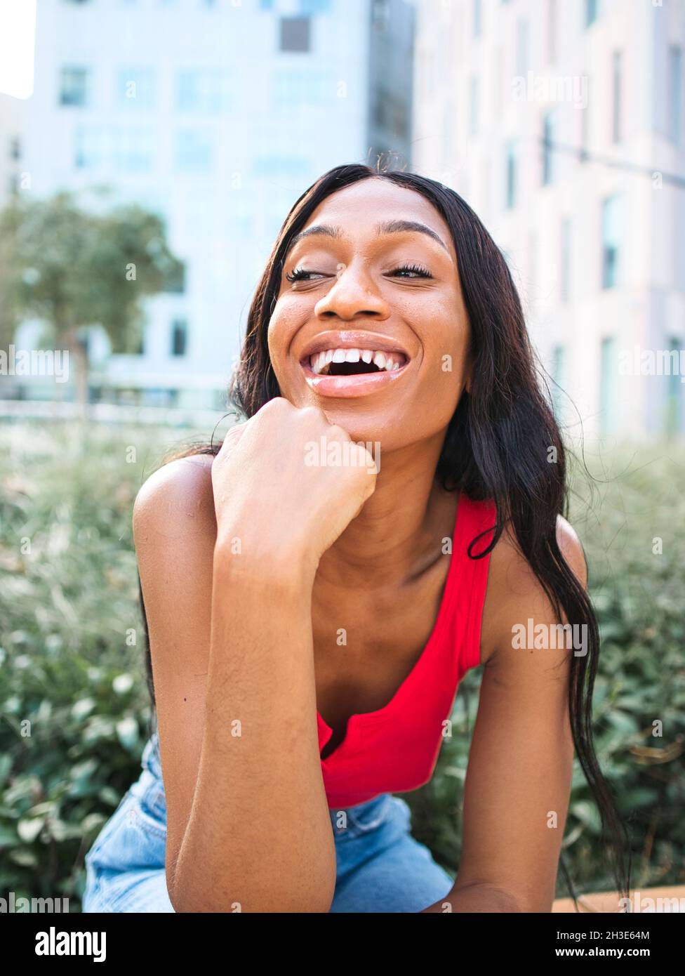 Happy African American femmina in abiti casual appoggiati a mano con sorriso e guardando via mentre si siede in strada vicino a cespugli verdi Foto Stock