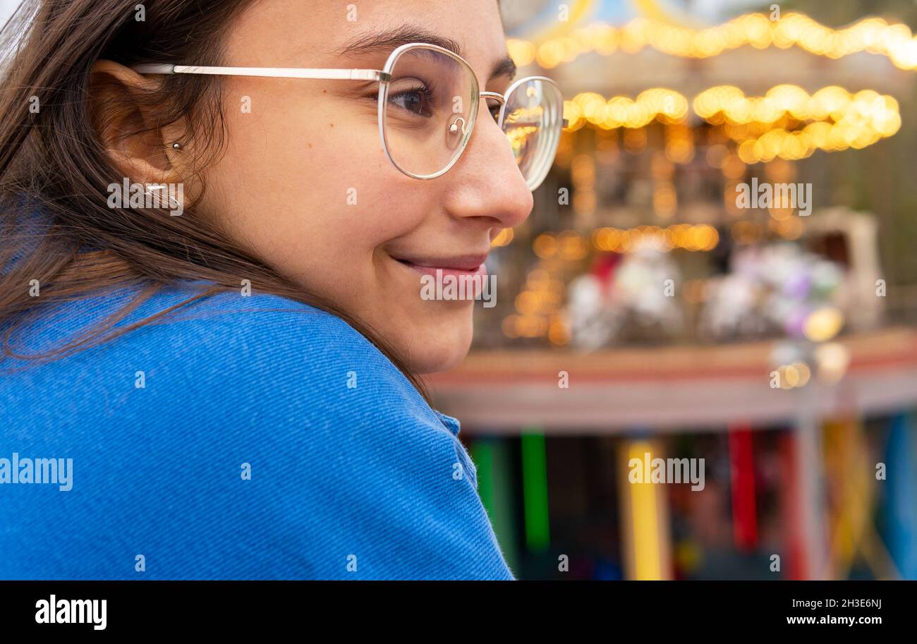 Giovane donna positiva in abiti casual e occhiali che guardano lontano mentre si trova vicino al carosello sullo sfondo della strada cittadina in luce del giorno Foto Stock