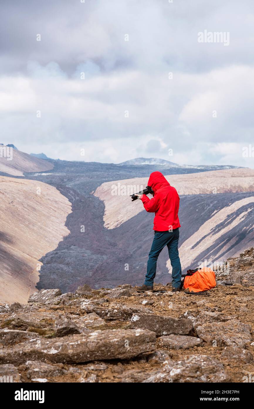 Vista laterale del fotografo maschile in outerwear in piedi sulla cima di una scogliera rocciosa vicino al vulcano attivo Fagradersfjall con lava nera in Islanda durante il giorno Foto Stock