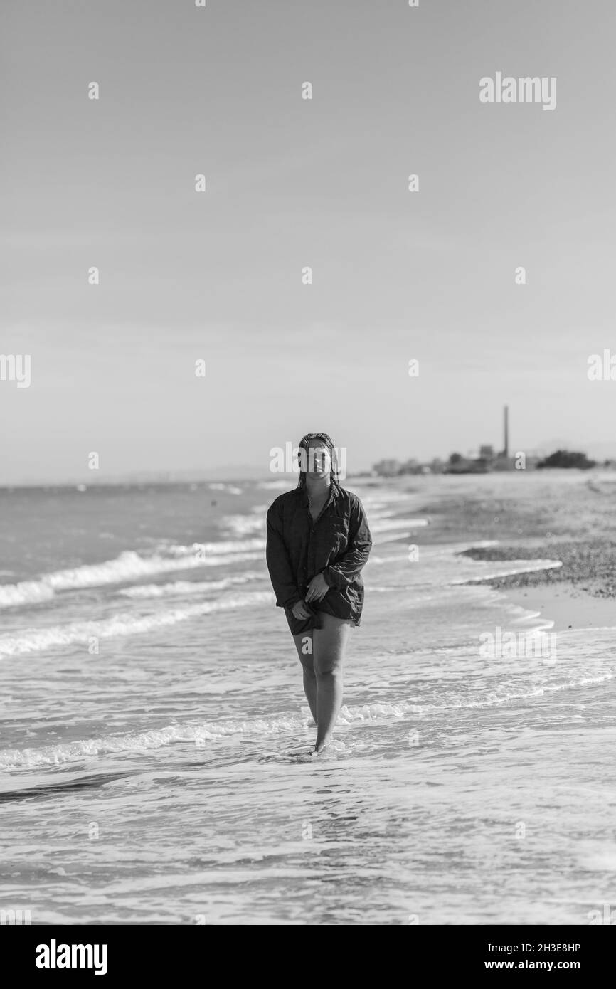 Bianco e nero di femmina a piedi nudi in camicia bagnata che cammina lungo la spiaggia sabbiosa vicino al mare ondeggiante in estate Foto Stock