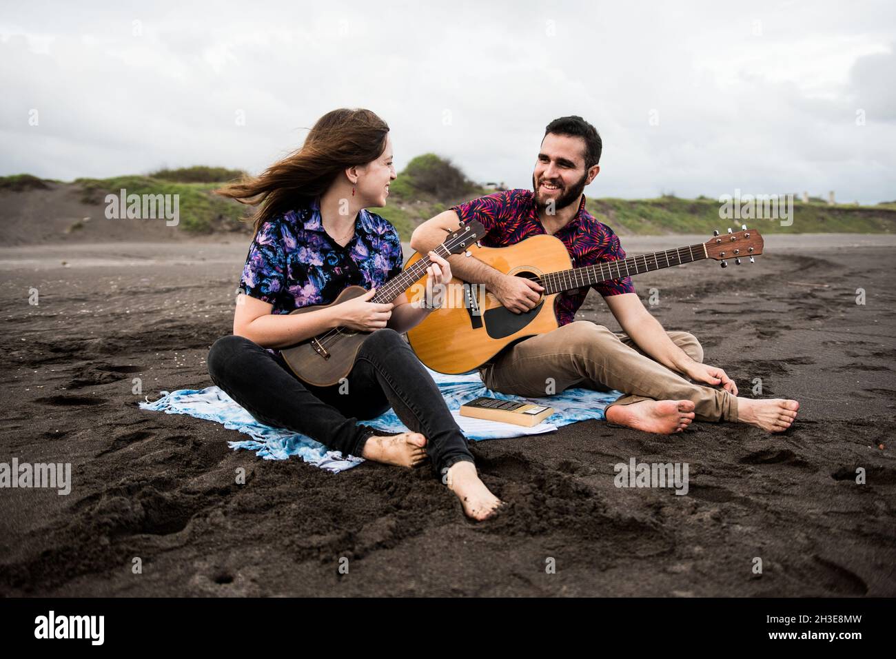 L'intera lunghezza di uomo sorridente suona la chitarra acustica con un amico positivo che suona l'ukulele mentre si siede sulla costa sabbiosa in natura in una giornata nuvolosa Foto Stock