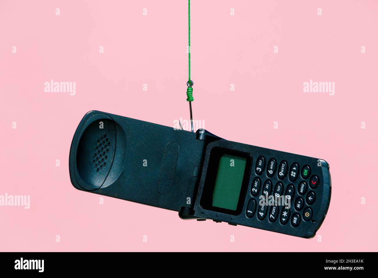 Vecchio telefono cellulare nero aperto flip appeso su gancio di metallo con corda verde su sfondo rosa in studio moderno chiaro Foto Stock