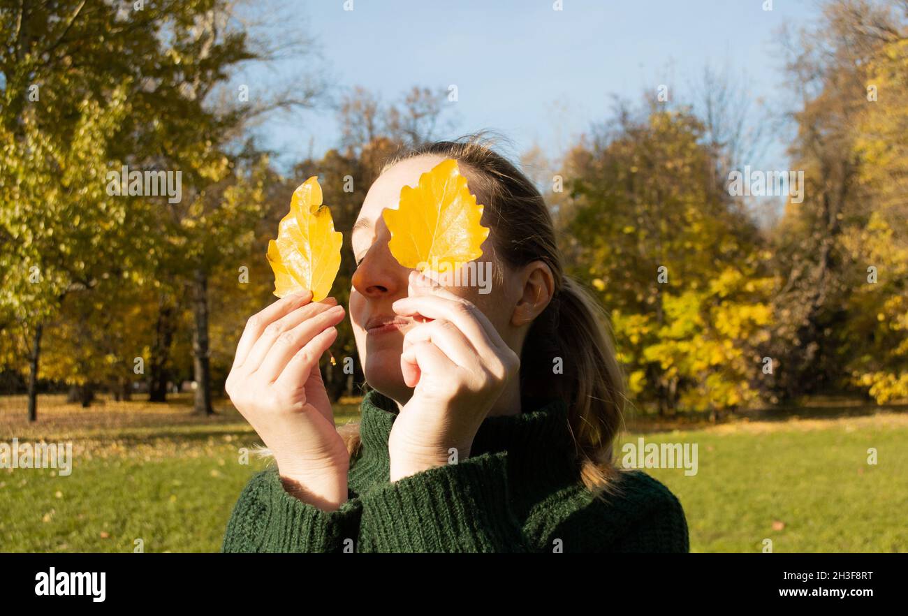 Ritratto di donna con gli occhi chiusi tiene due foglie gialle, mani ravvicinate, sfondo alberi autunnali. Foto Stock