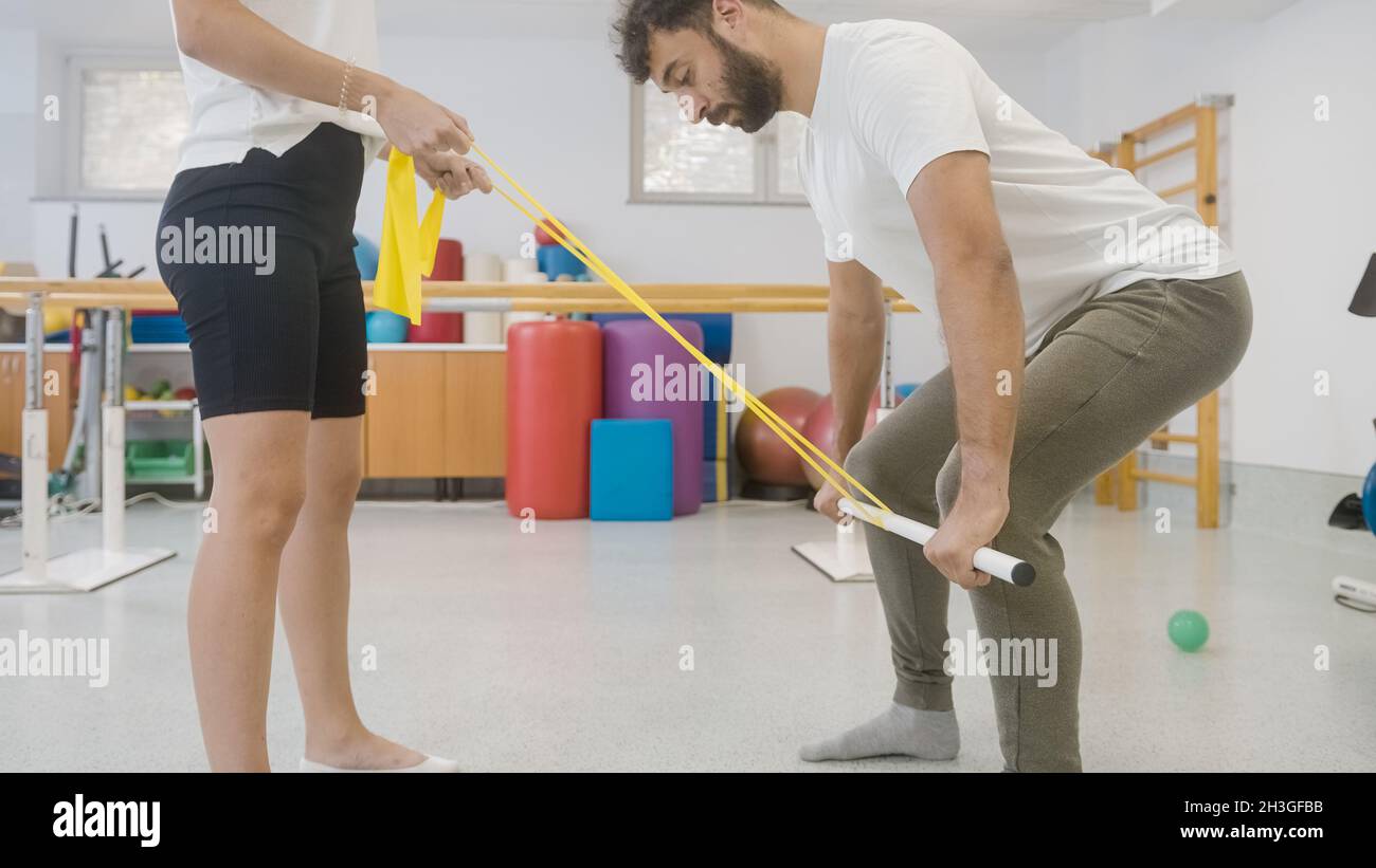 Coppia durante la terapia di riabilitazione presso la clinica. Uomo che fa un allenamento completo con la barra medica e le fasce di resistenza. Foto Stock