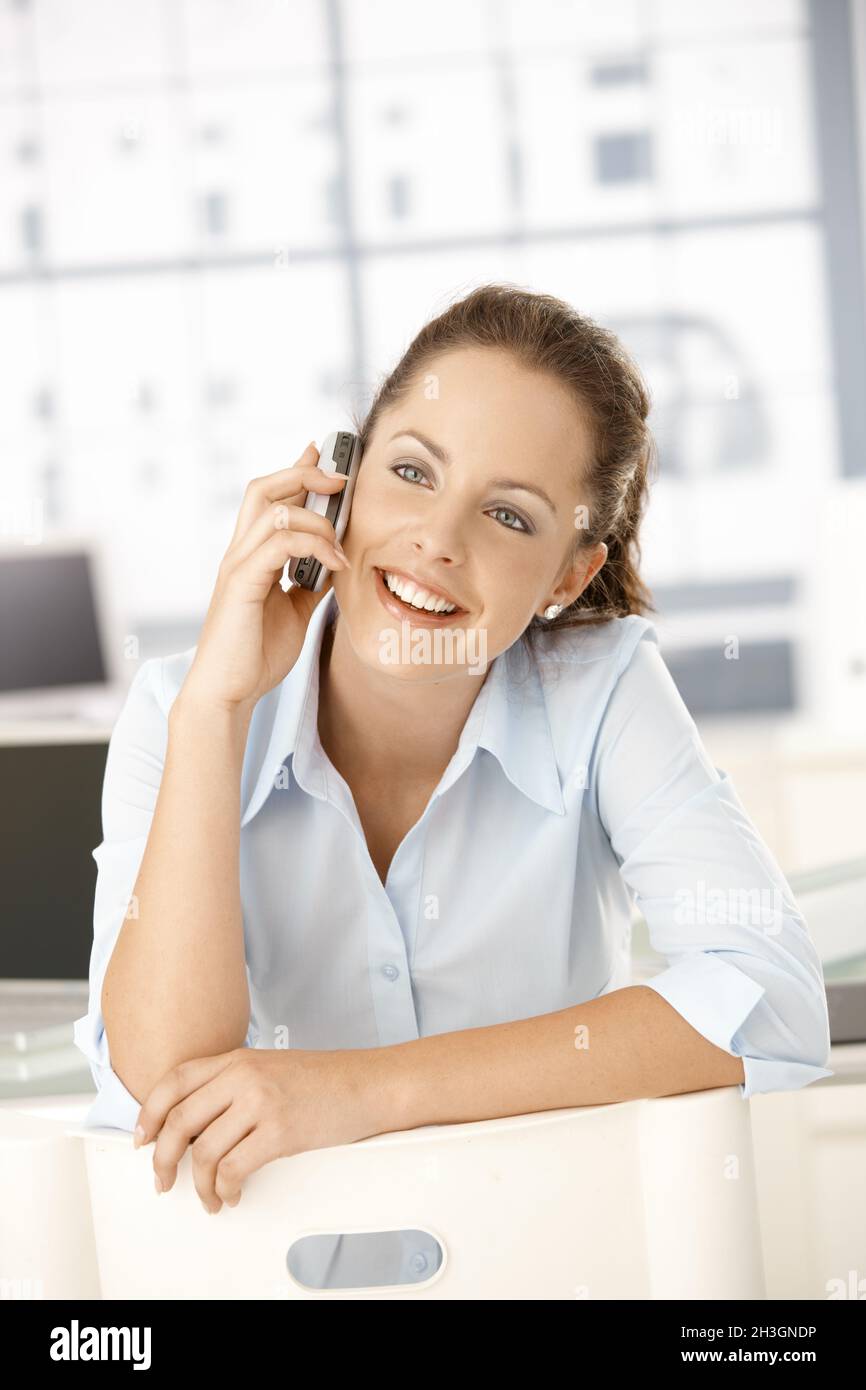 Giovane femmina che chiacchiera su cellulare seduto in ufficio Foto Stock