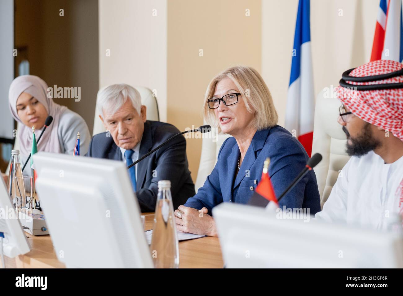 Donna caucasica matura e fiduciosa in occhiali seduti a tavola con altri politici e utilizzando il microfono durante la conferenza politica Foto Stock