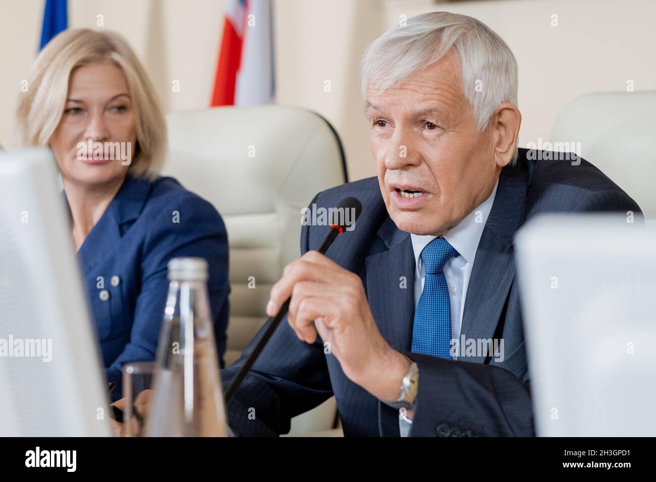 Politico caucasico senior in costume formale seduto al tavolo della conferenza e utilizzando il microfono mentre si parla di problema globale Foto Stock