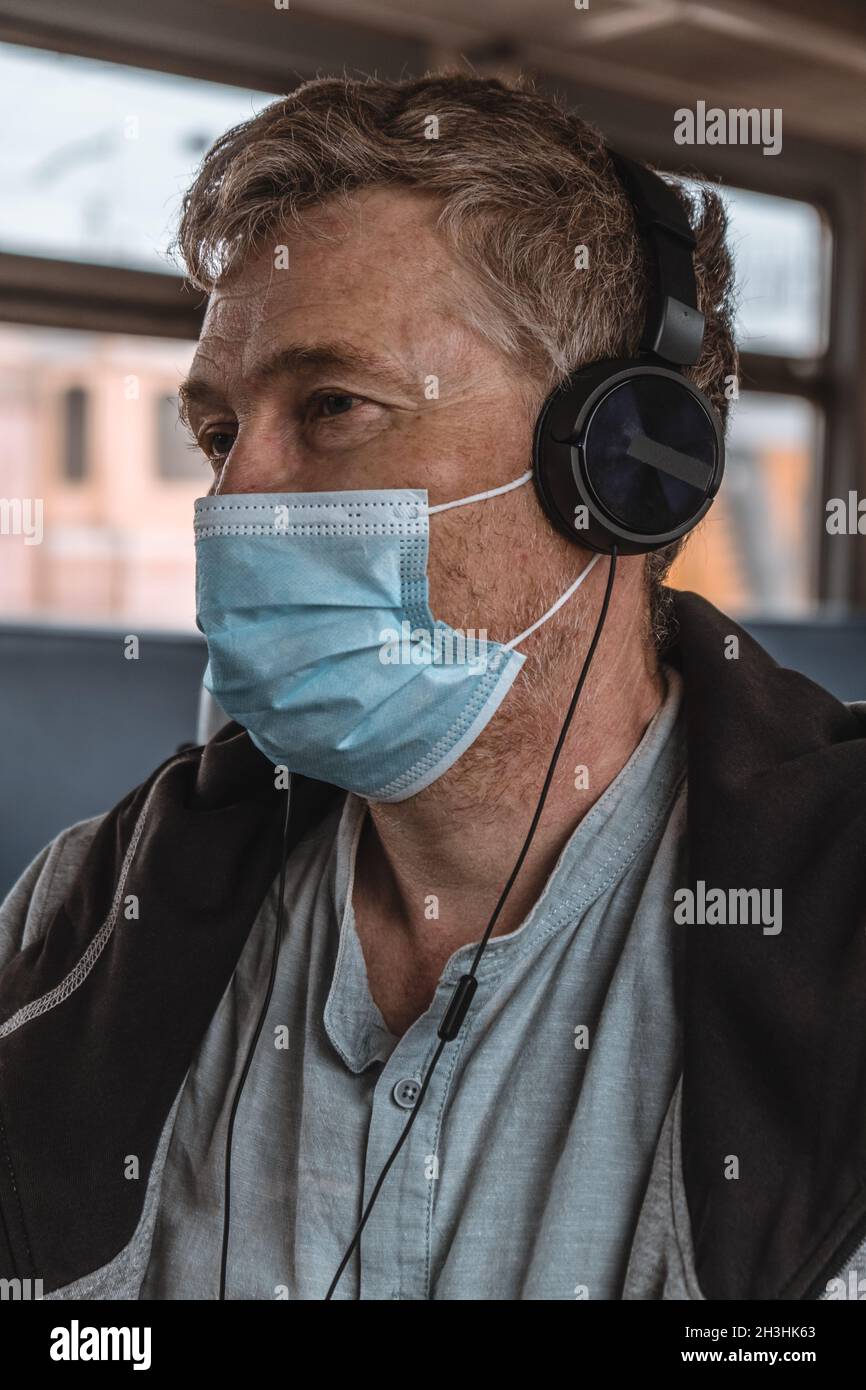 Ritratto di un uomo adulto serio che indossa le cuffie con una maschera medica protettiva sul suo viso nei trasporti pubblici vicino alla finestra. Distanza sociale. Foto Stock