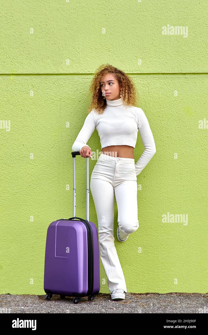 Bella ragazza giovane con capelli ricci che aspetta felicemente con una valigia viola su sfondo verde Foto Stock