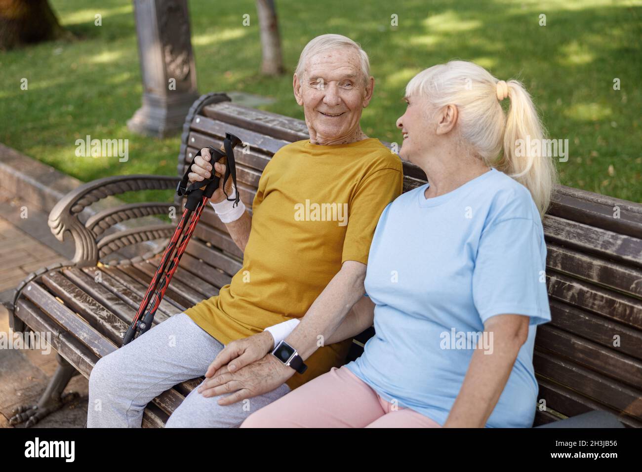 Uomo anziano felice con le aste per nordic walking e la moglie si siedono sulla panchina nel parco Foto Stock