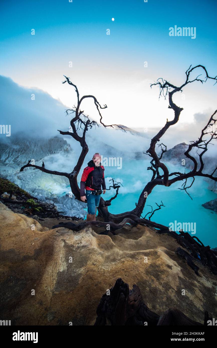 Uomo viaggiatore in piedi vicino albero morto sul bordo del vulcano Ijen del cratere con cielo colorato al mattino Foto Stock