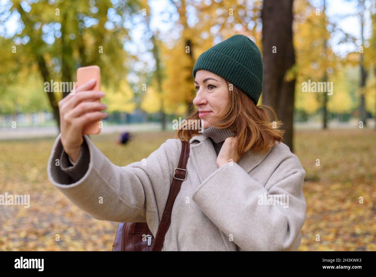 Donna che fissa i capelli o il look e guarda allo schermo del telefono attraverso la fotocamera anteriore come specchio al parco autunnale Foto Stock