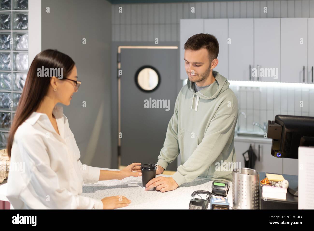 Uomo in felpa con cappuccio che dà la bevanda calda alla donna asiatica mentre lavora nella moderna caffetteria Foto Stock