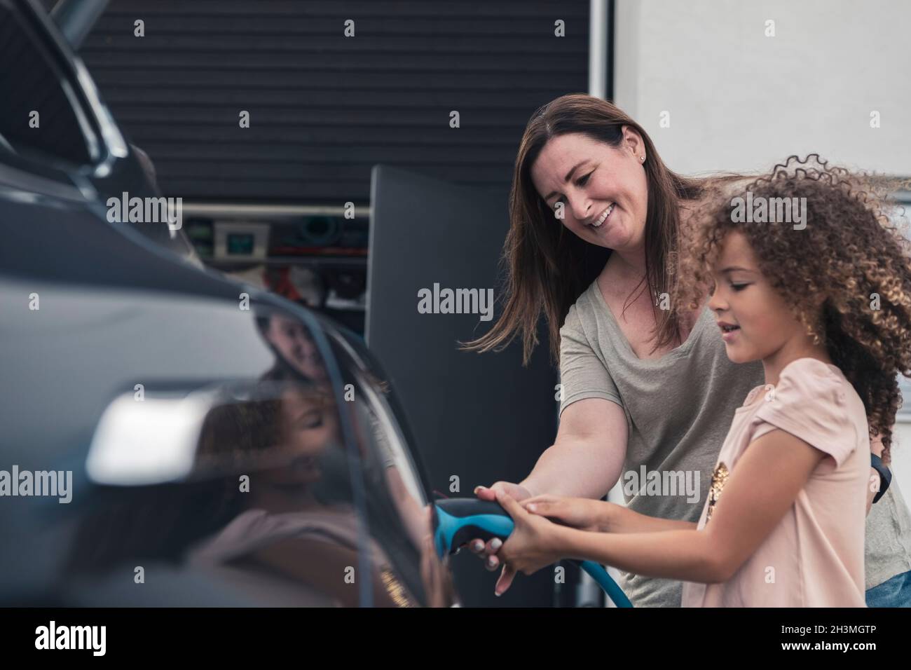 Madre sorridente che aiuta la figlia a caricare l'auto elettrica nel cortile anteriore Foto Stock