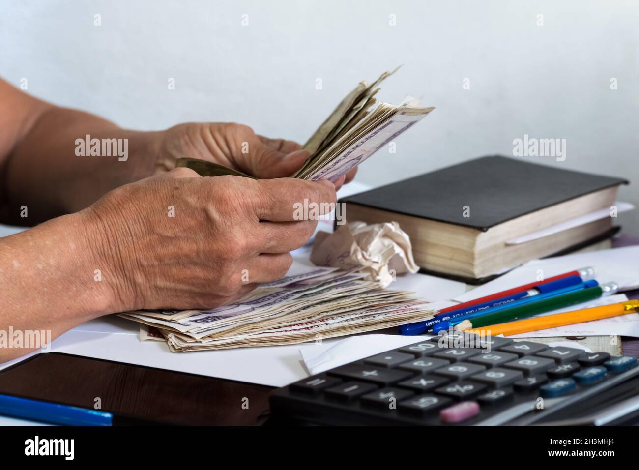 Primo piano di mani femminili senior contando denaro su una scrivania. C'è un libro chiuso, matite, un telefono cellulare e una calcolatrice sulla scrivania. Foto Stock