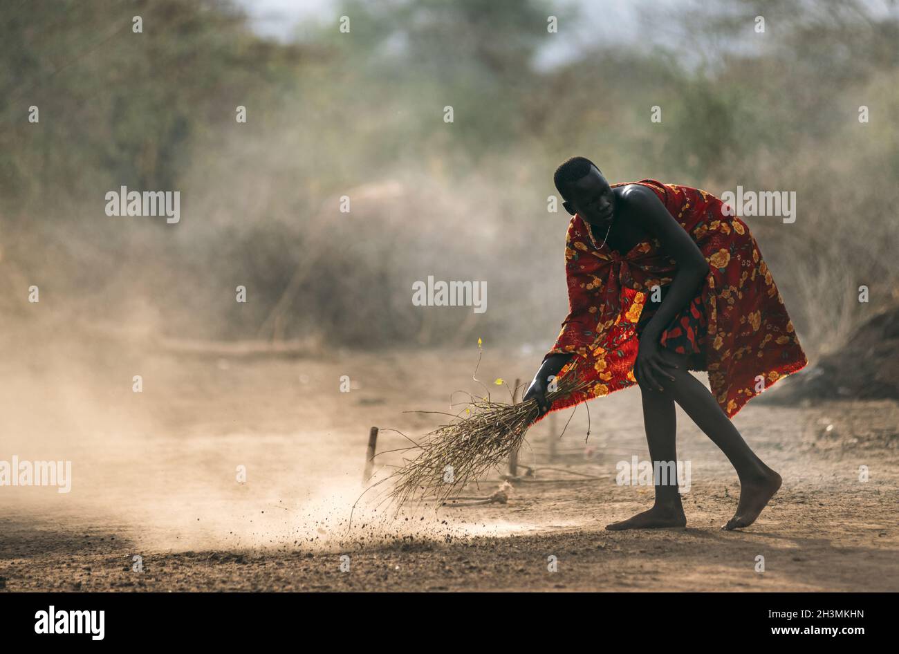 TRIBÙ MUNDARI, SUDAN DEL SUD - 11 MARZO 2020: Uomo in abito luminoso spazzando via terra polverosa con ramoscelli e guardando via a Mundari Foto Stock