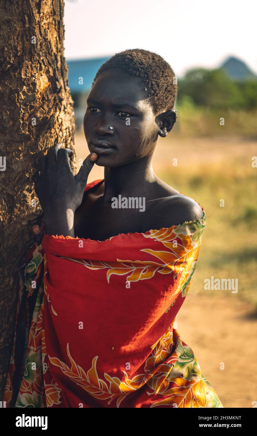 TRIBÙ DI BOYA, SUDAN DEL SUD - 10 MARZO 2020: Giovane donna in abito colorato che tocca il labbro e guarda via mentre si appoggia sulla savana Foto Stock