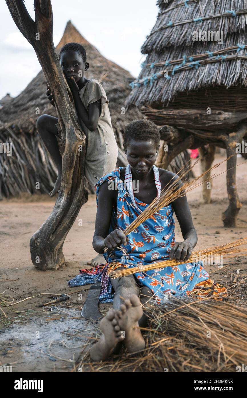 Tribù TOPOSA, SUDAN DEL SUD - 12 MARZO 2020: Donna seduta su terra sporca vicino a capanna e capretto e prodotto di tessitura da paglia nel male Foto Stock