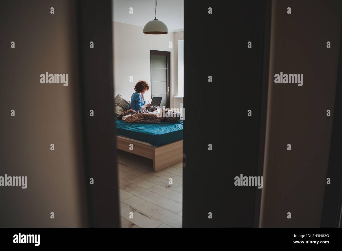 Donna con capelli ricci a partire la mattina prendendo lezioni online sul portatile dal letto ancora sudicio. Formazione online. Vista dalla porta parzialmente aperta Foto Stock