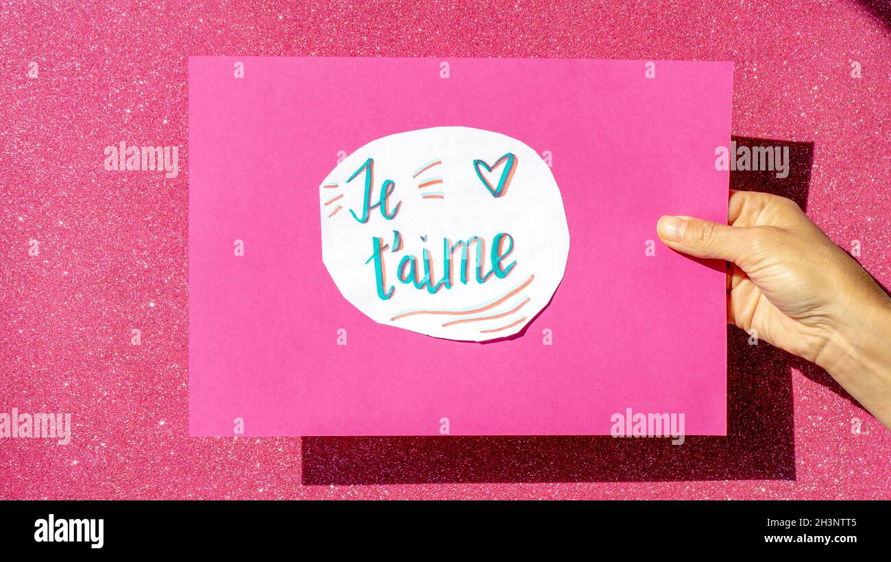 insegna manoscritta su carta bianca incollata su un cartone rosa tenuto da una mano con le parole je t' aime Foto Stock