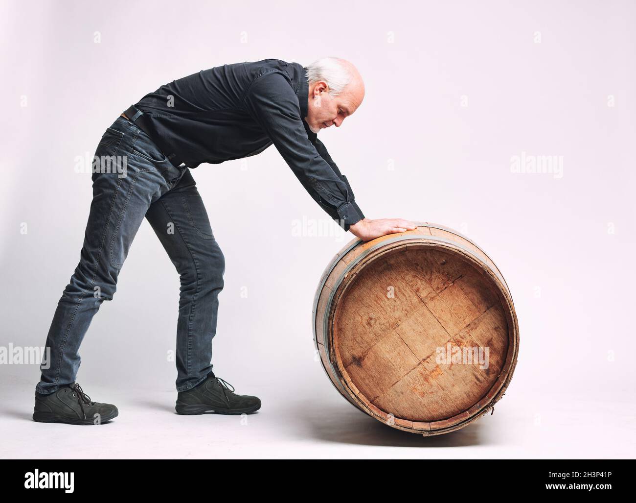 Uomo anziano che rotola un vecchio vino di quercia o barile di birra di fronte a lui lungo il pavimento in una vista laterale completa su bianco Foto Stock