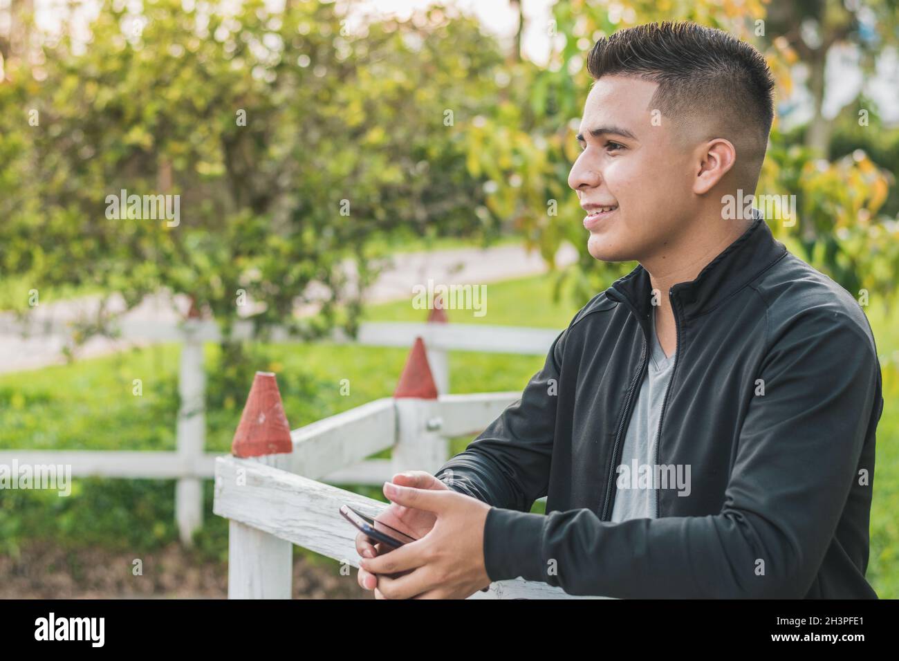 intraprendente giovane uomo accanto alla recinzione di legno bianco con il suo telefono cellulare in mano guardando in su, felice pensando ai suoi successi, con una bellezza Foto Stock