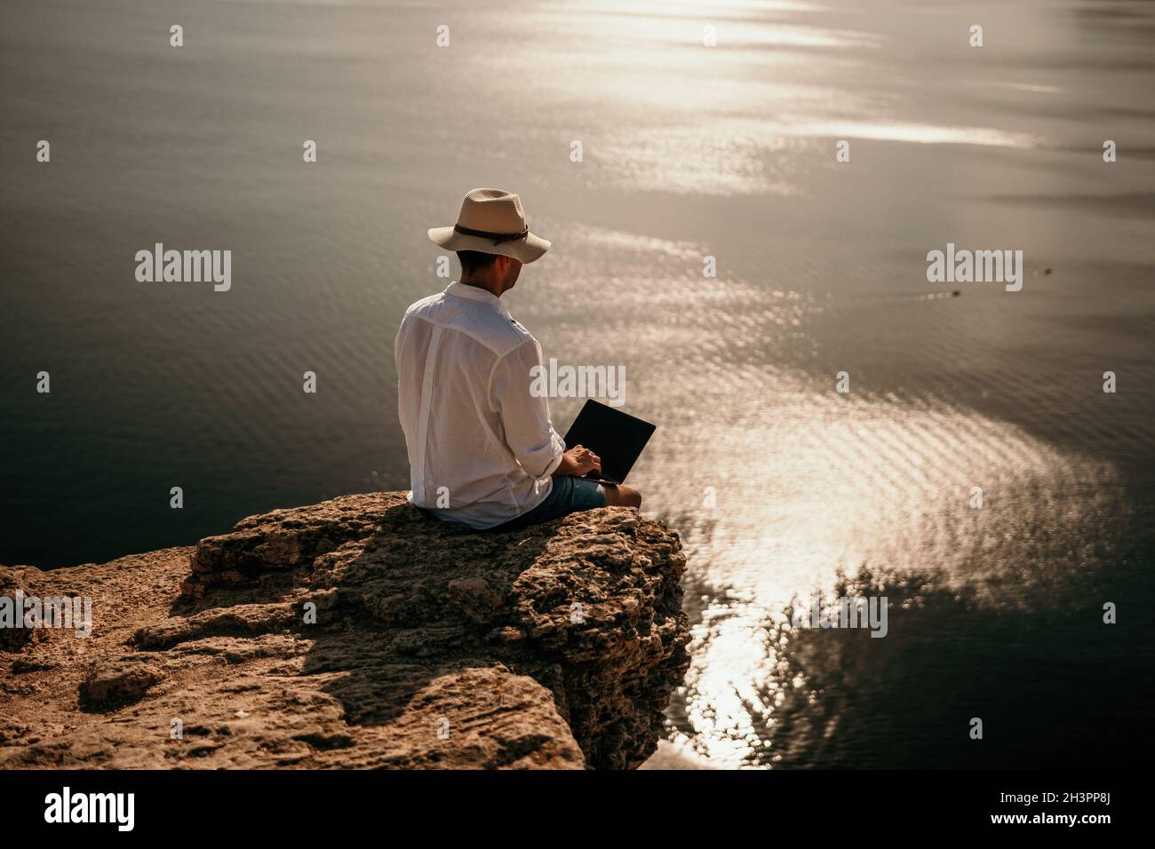 Digital nomad, uomo nel cappello, un uomo d'affari con un laptop siede sulle rocce vicino al mare durante il tramonto, effettua una transazione di affari in linea da a. Foto Stock