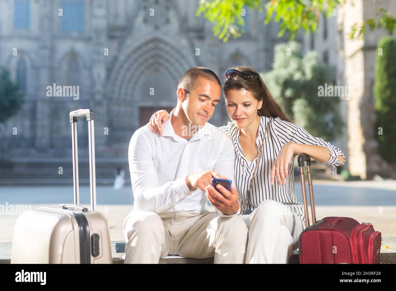 Coppia di viaggiatori seduti su panca, prenotazione di alloggi con telefono Foto Stock