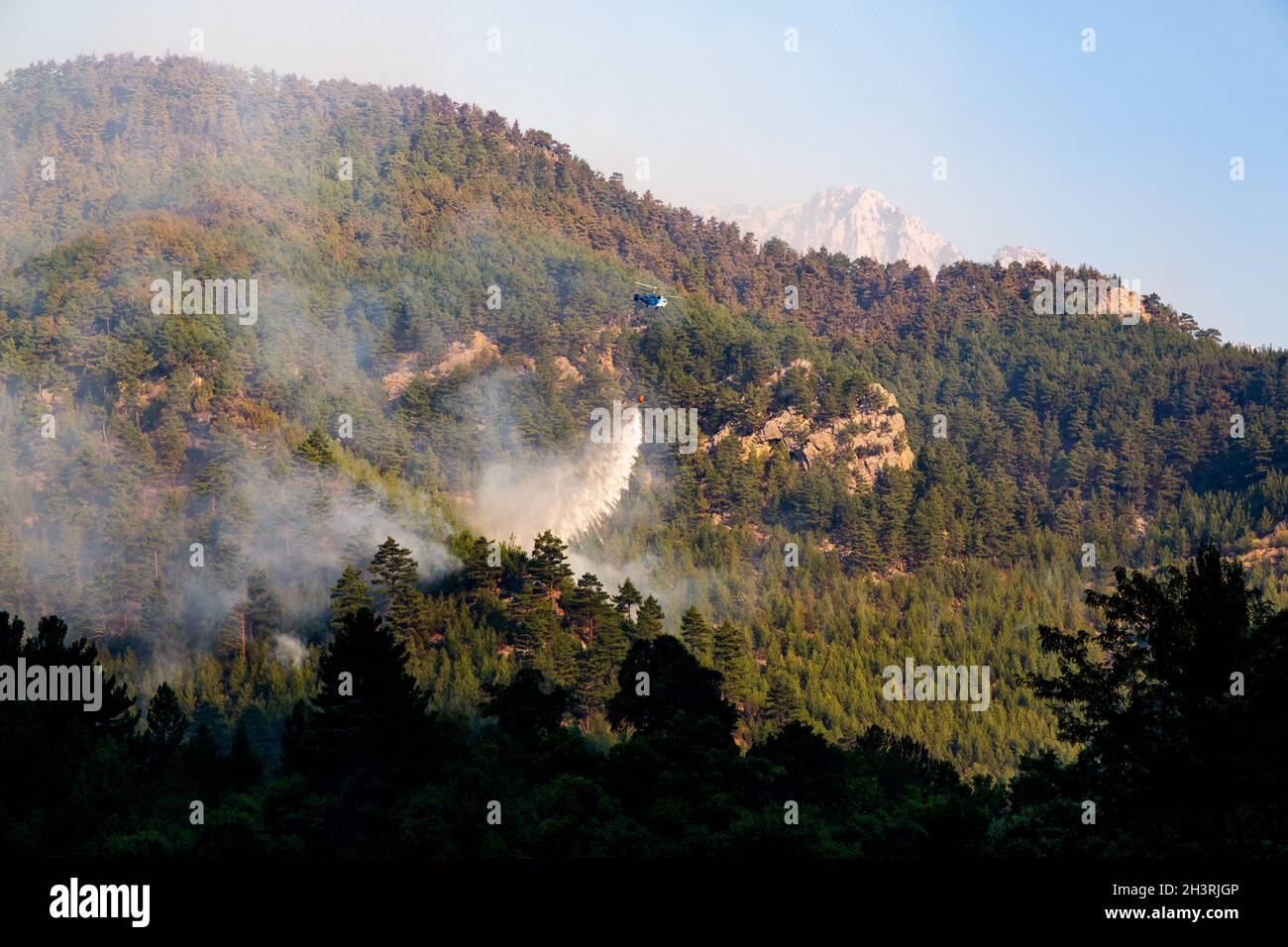 L'elicottero Wildfire fa cadere l'acqua su un fuoco in un terreno roccioso e ripido con un secchio d'acqua. Le montagne sono coperte da fitte pinete. Foto Stock