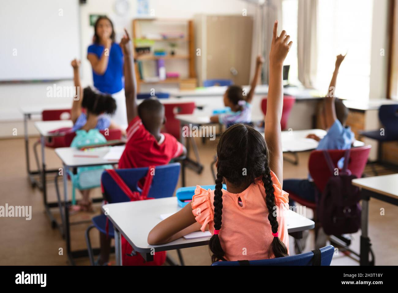 Vista posteriore degli studenti che alzano le mani mentre si siedono sulla scrivania in classe a scuola Foto Stock